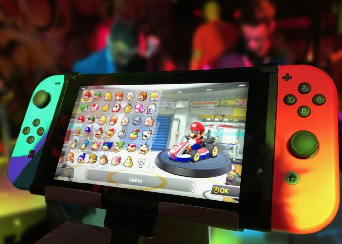 Close-up of a Nintendo Switch showing Mario Kart selection screen, gaming atmosphere in the background.