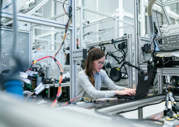 A female engineer focuses on her laptop amidst advanced tech equipment in a lab.
